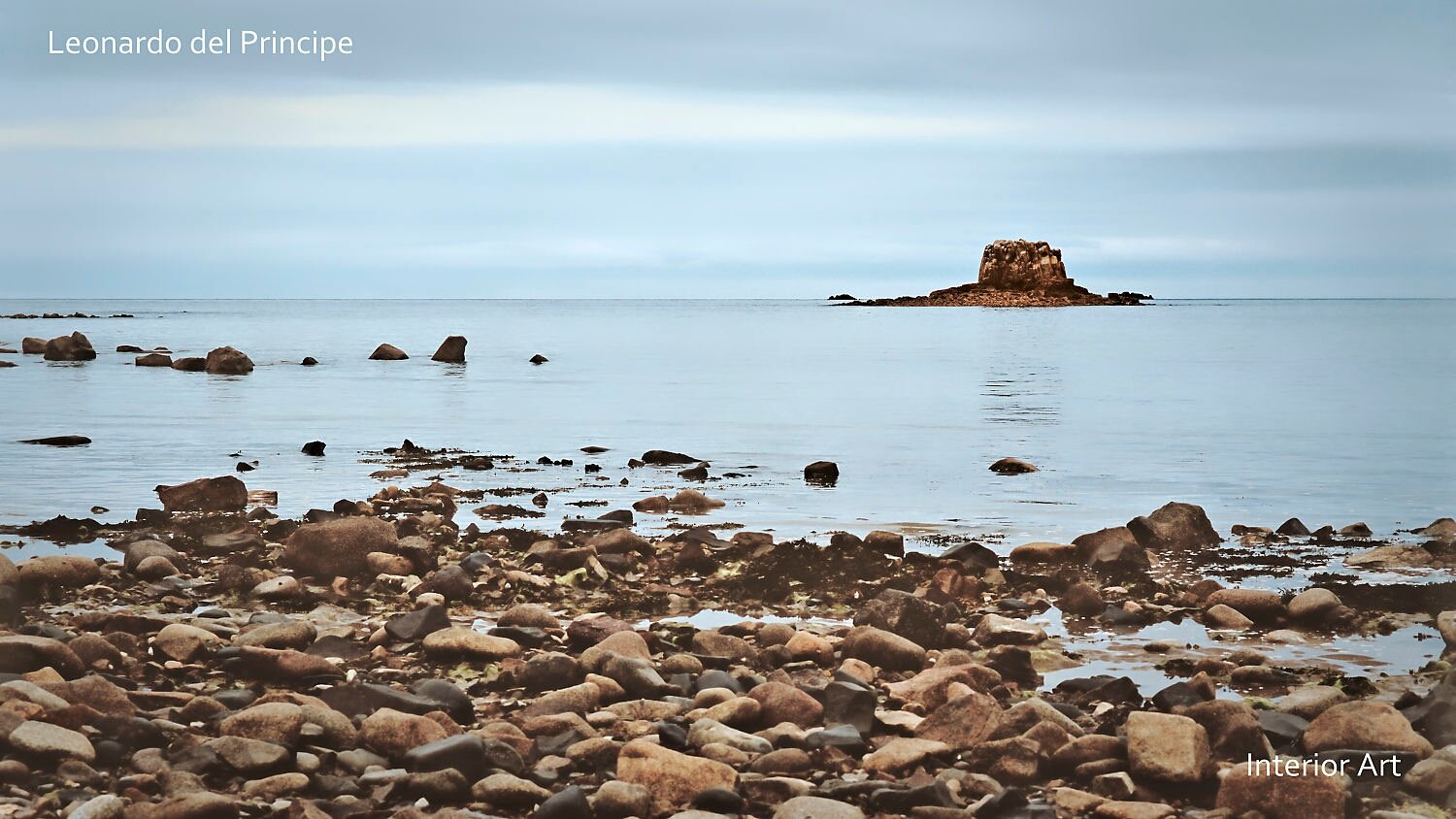 MORG03 Seascape with rocky shore in foreground. A small, isolated rock formation emerges from the serene blue sea under a cloudy sky. Natural monument.